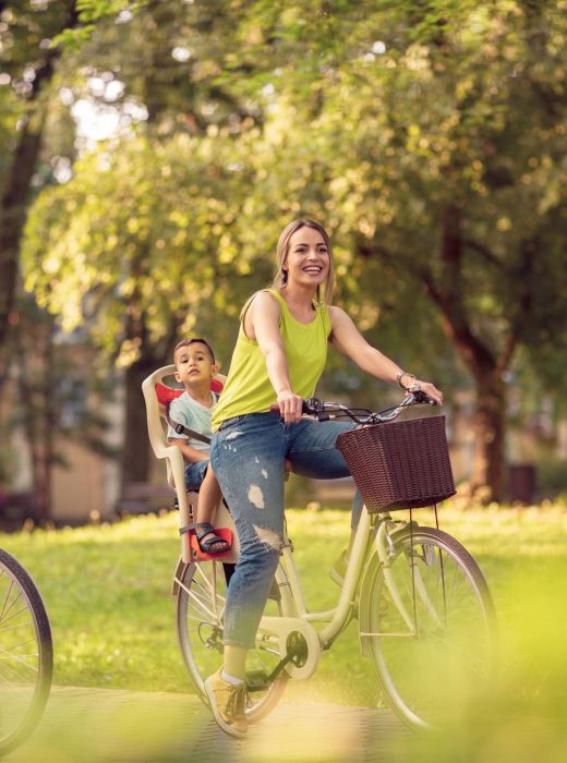A family rides bikes through a sunny, green park; a child sits in a seat behind the smiling woman.
