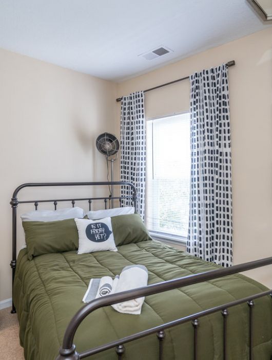 A neatly made bed with green bedding in a bright bedroom with patterned curtains and geometric mirrors.