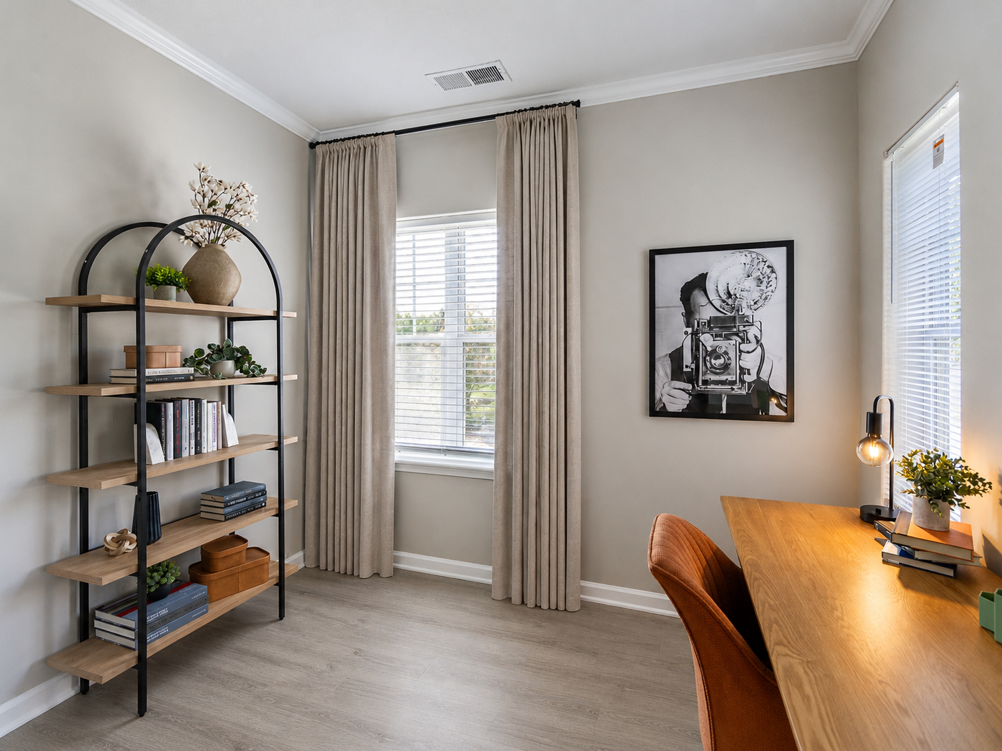 A home office with a wooden desk, orange chair, bookshelf, large window, beige curtains, and a black-and-white camera art print on the wall.