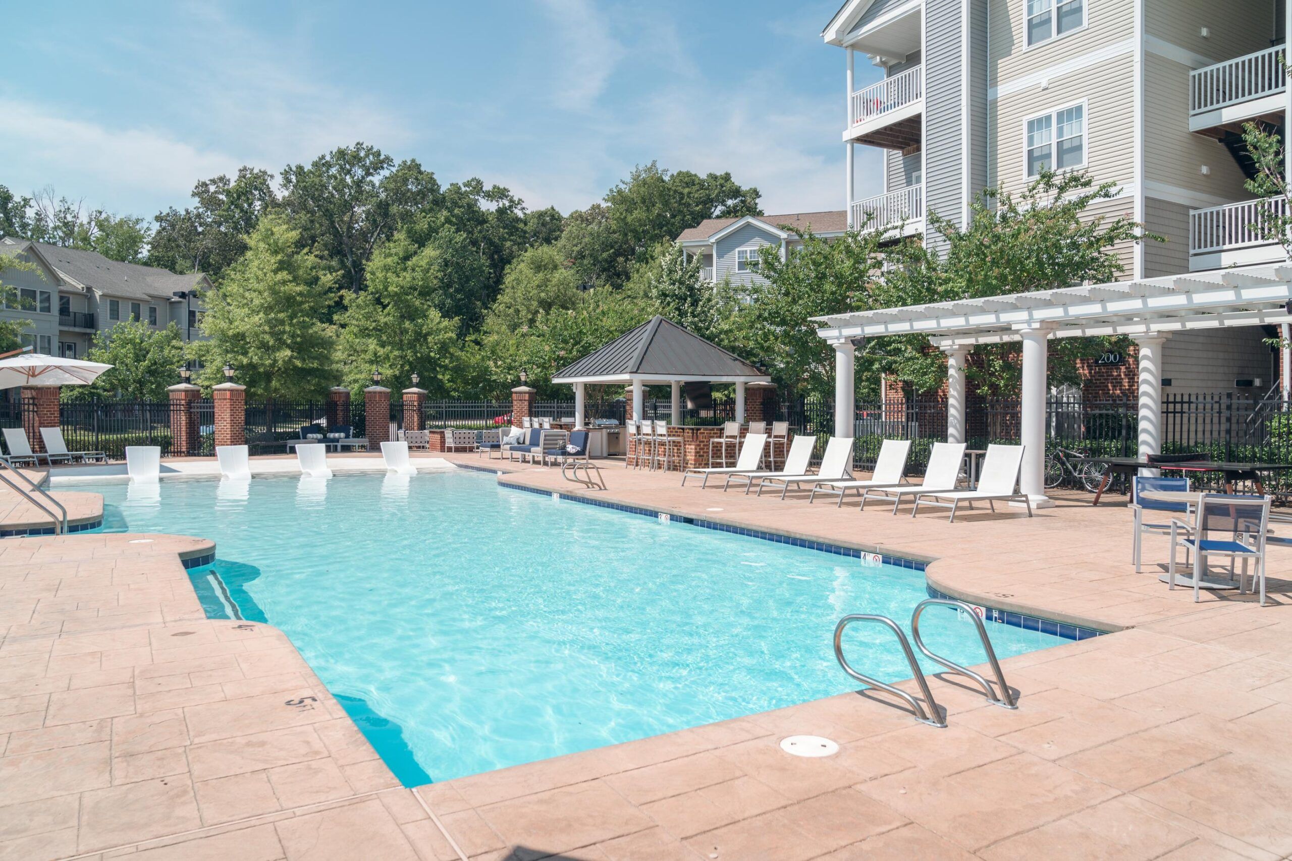 Outdoor swimming pool with lounge chairs, pergola, and trees near apartment buildings on a sunny day.