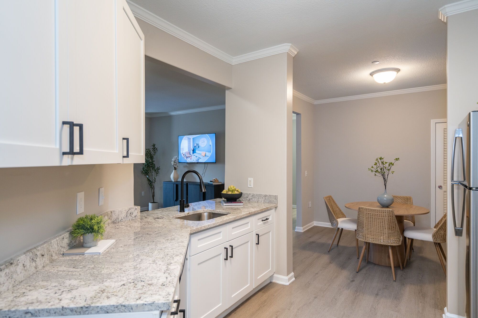 A white kitchen with speckled countertops and a black faucet, opening into a dining area with a round wooden table and wicker chairs.
