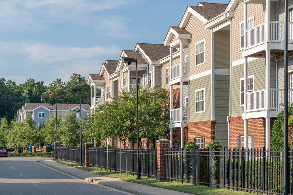 Row of modern apartment buildings with balconies, trees, and a black metal fence along a quiet street.