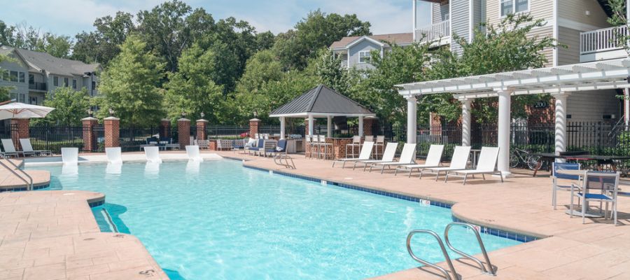 Sunny outdoor pool with lounge chairs, a shaded pavilion, and apartment buildings in the background.